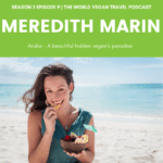 A woman in a blue shirt eating fruit on a beach in Aruba with the text The World Vegan Travel Podcast - Meredith Marin Aruba an excellent vegan destination
