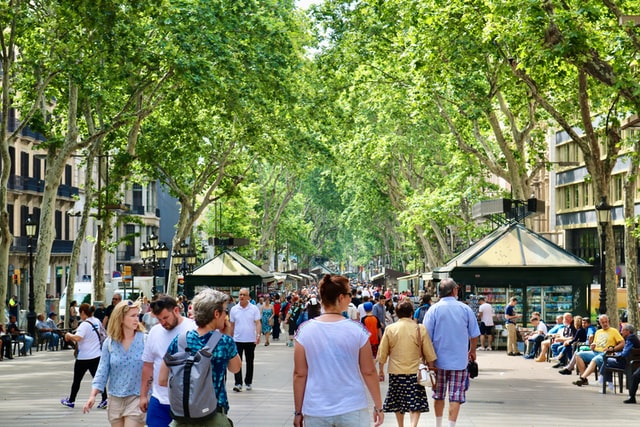 Vegan Walking City food tour World Vegan travel Barcelona La ramblas Many people in Barcelona. Walking up and down La Rambas in Barcelona Spain. There are lots of overhanging trees and tables at the side with many people relaxing and enjoying the summer.