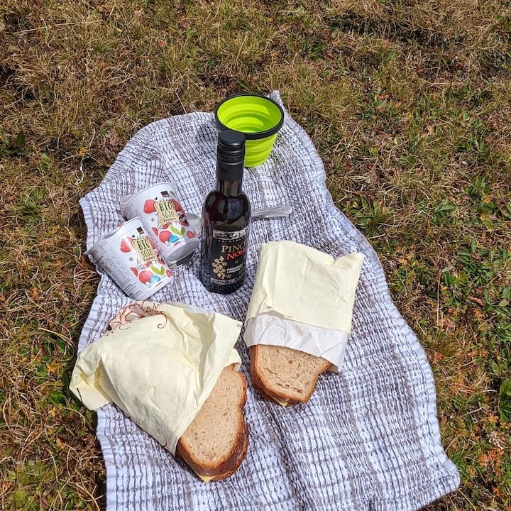 A tea towel being used as a picnic blanket with two sandwiches, yogurts and a small glass of wine.