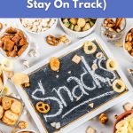 A flat-lay image of bowls filled with different vegan snacks like popcorn, pretzels, and crackers surrounding a chalkboard that says “Snacks.”