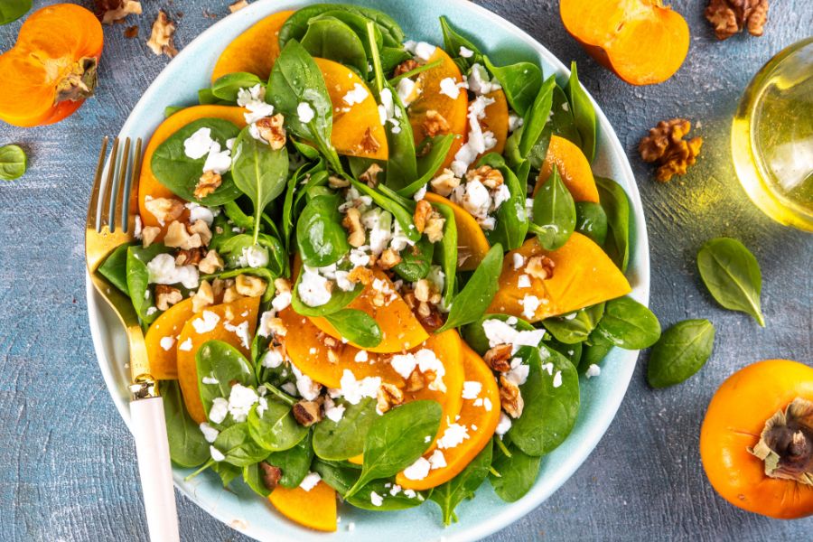 Bright salad with persimmons, spinach, walnuts, and vegan cheese on a blue plate, World Vegan Travel.