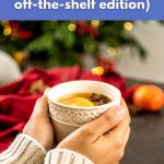Hands holding a warm cup of vegan holiday drink with slices of orange and star anise, in front of a festive Christmas background.