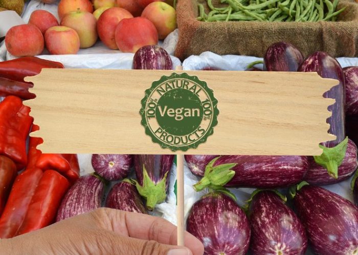 Hand holding a “Vegan Products” sign over fresh vegetables and fruit with the text “Best Vegan Products at Costco.”