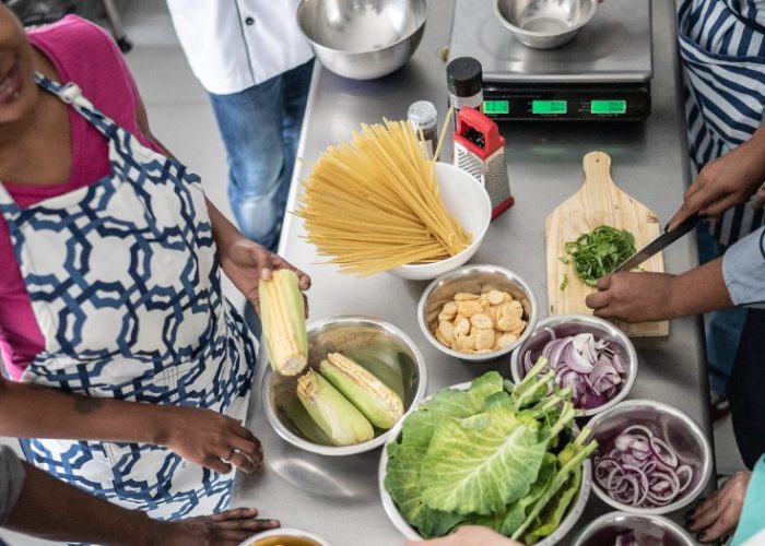 People preparing fresh vegetables and pasta in a cooking with text overlay “The Best Vegan Online Courses to Gift for the Holidays” from World Vegan Travel.