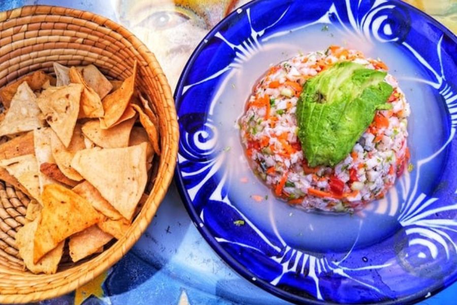 A plate of colorful ceviche topped with avocado, served with a basket of crispy tortilla chips.