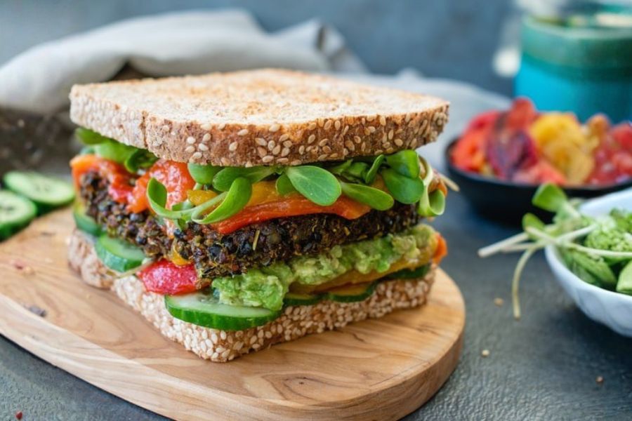 A tall vegan sandwich filled with veggies, greens, and spreads on seeded bread.