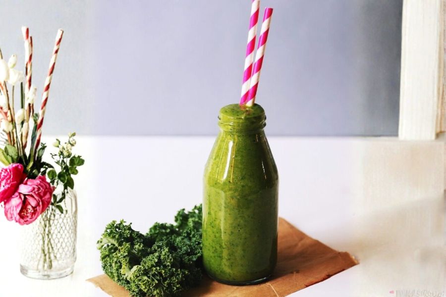 Green vegan smoothie bottle with pink straws next to kale and flowers on a wooden surface.