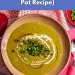 A bowl of creamy vegan split pea soup topped with herbs and peas, served on a table with a pink cloth.