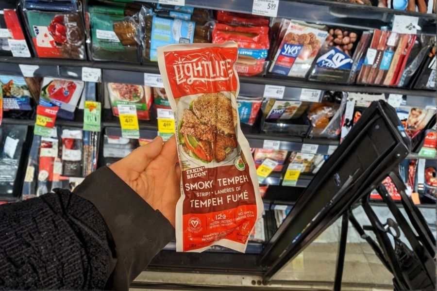 A hand holding a packet of smoky tempeh in front of a supermarket fridge filled with vegan products.
