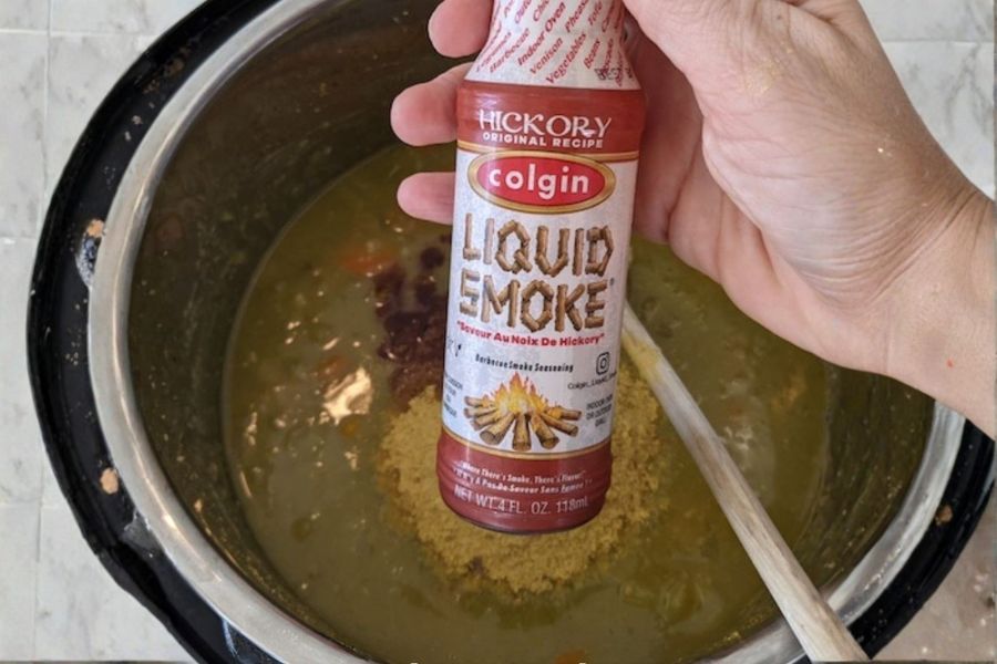 Hand holding a bottle of liquid smoke over a pot of vegan split pea soup.