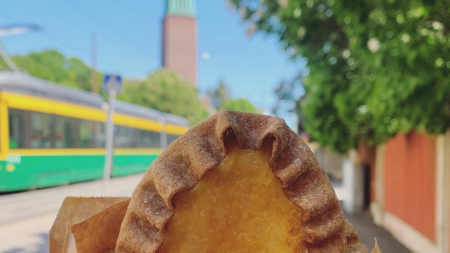 Hand holding a vegan Karelian pie with Helsinki tram and church in background.