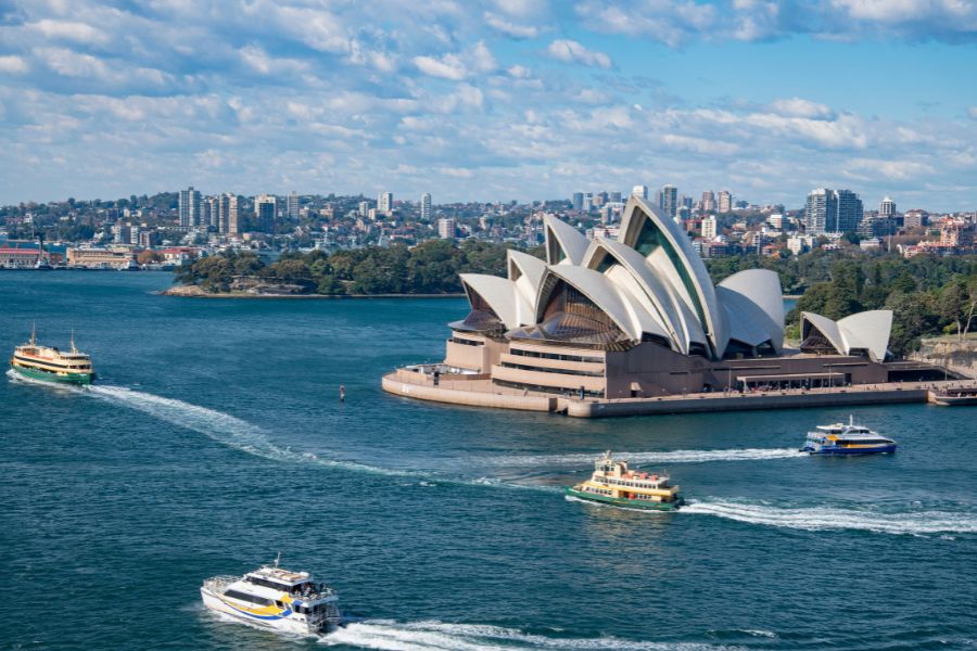 Boats and ferries sailing past the Sydney Opera House on a sunny day in Australia.