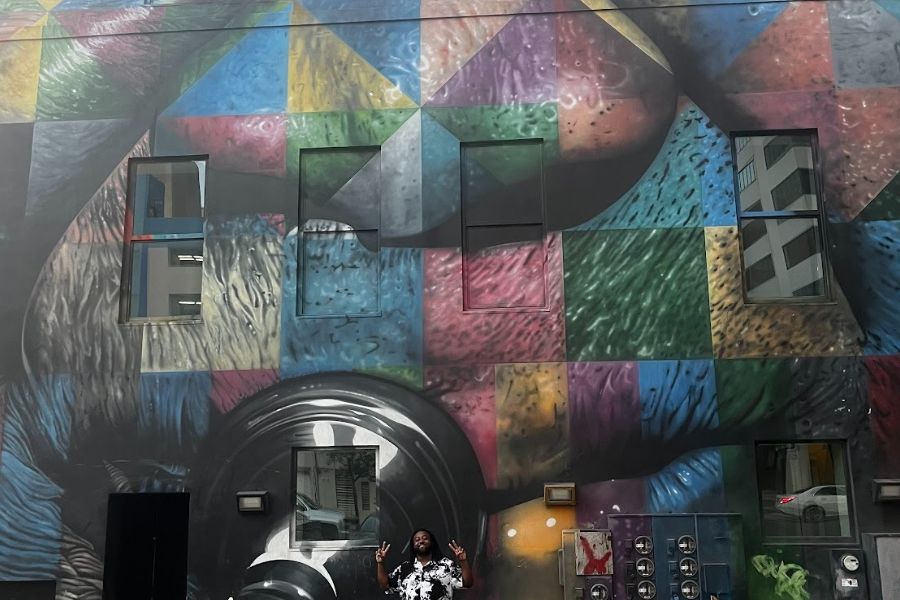 Original Simmons poses in front of a colorful mural, blending travel, art, and self-expression.