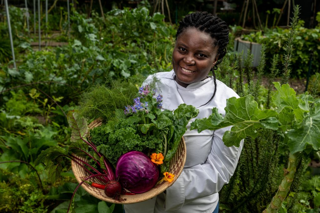 Jane Nshuti holding a basket of fresh vegetables in a garden, representing African plant-based cuisine