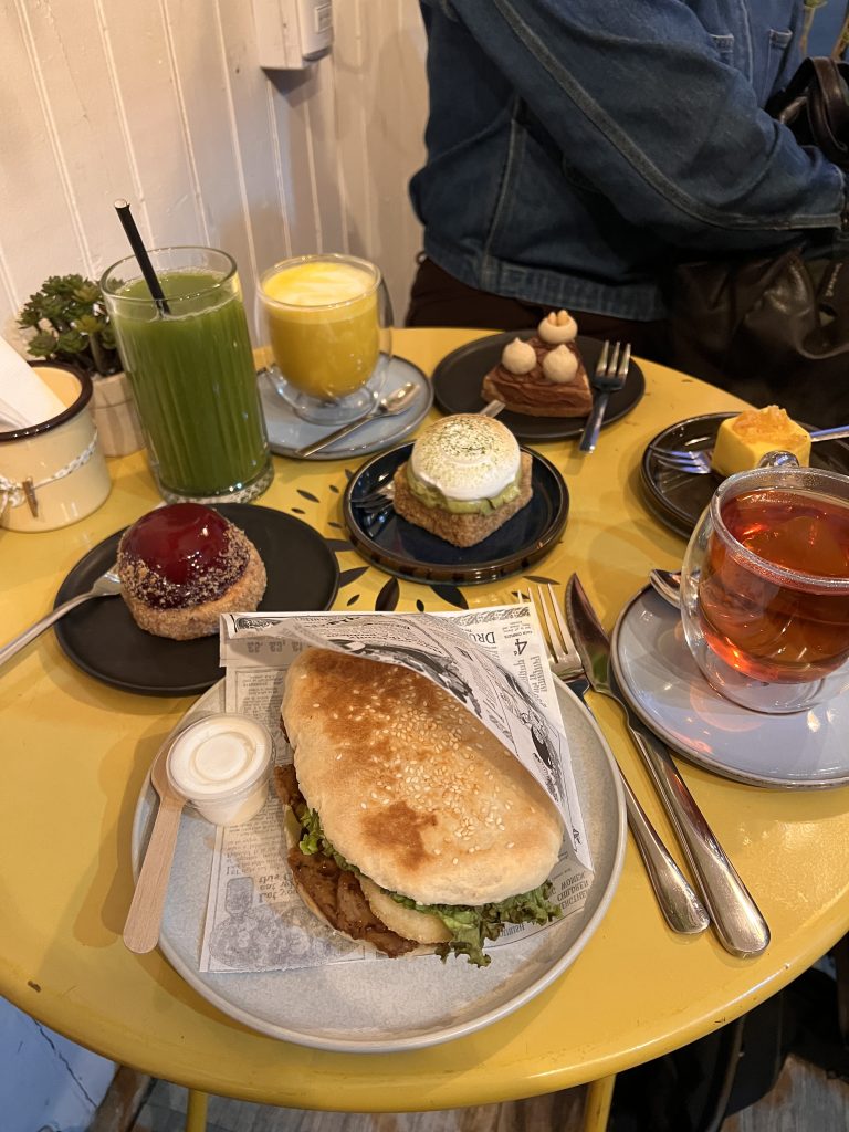 A vegan sandwich, assorted pastries, tea, and colorful drinks arranged on a yellow café table.