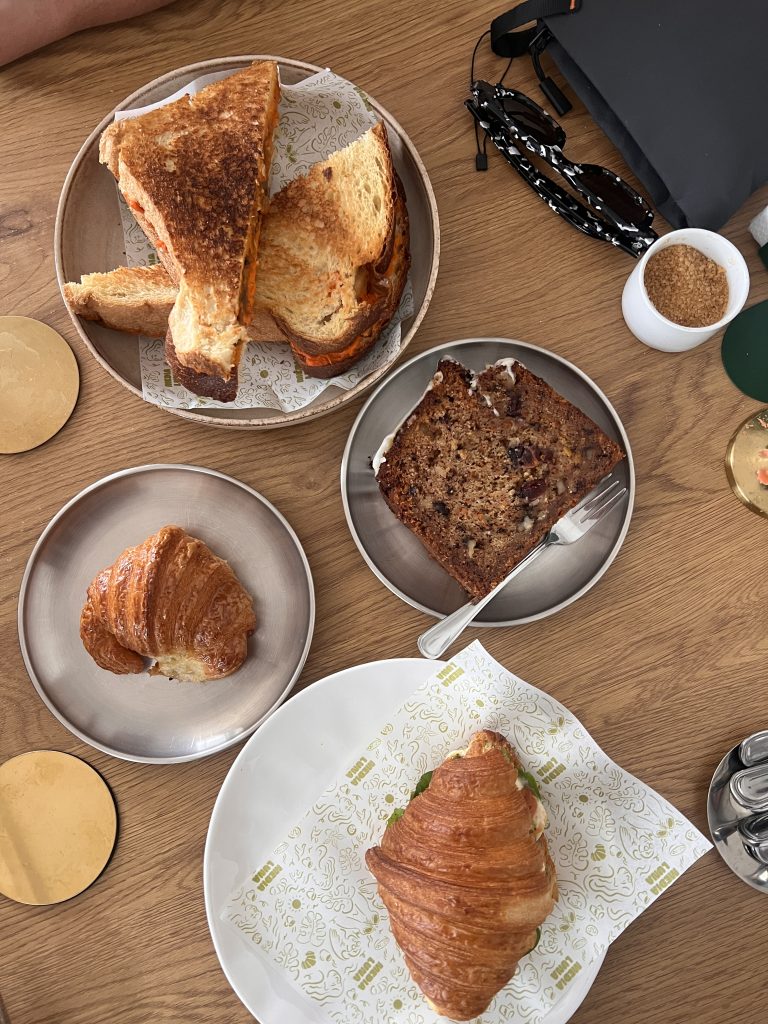 Toasted vegan sandwiches, two croissants, and a slice of cake arranged on a wooden café table.