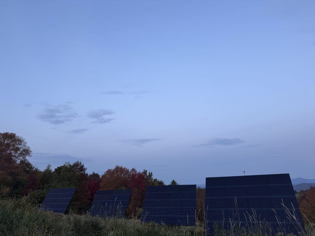 Four solar panel arrays in a grassy field at Balance.Farm, with autumn trees and distant mountains.