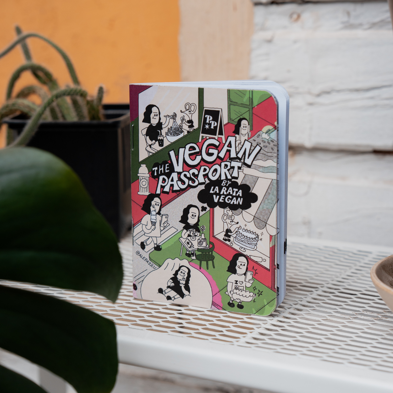 The illustrated cover of The Vegan Passport by La Rata Vegan, standing on a white table beside plants.